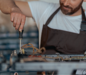 A close-up lifestyle shot of a man wearing a white t-shirt and a brown apron, working on a espresso machine repair task at a workbench.